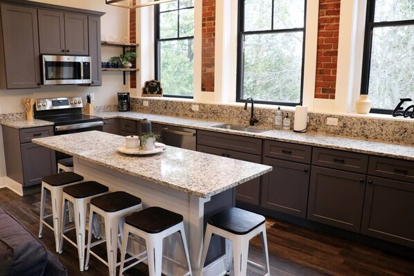 A modern kitchen with granite countertops, a central island with bar stools, stainless steel appliances, and large windows with a view of trees.