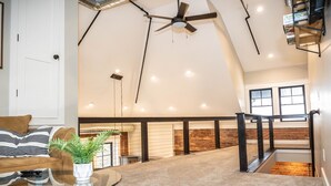 A spacious loft with a high ceiling, exposed brick walls, a modern ceiling fan, and a staircase leading to an upper level. - The Sunset View Penthouse Loft (Boyne City)