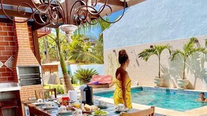 An outdoor dining area with a pool, wooden furniture, and a brick oven. - Villa Lulu Buzios (Búzios)