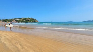 A beach with a sandy shore, a person carrying a surfboard, and a hill with buildings in the background. - Villa Lulu Buzios (Búzios)