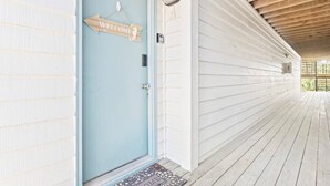 En lyseblå dør med et 'WELCOME'-skilt, en træterrasse og en hvid facade.