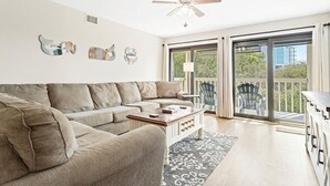 En moderne stue med en beige hjørnesofa, et træsofa bord og en stor skydedør, der fører ud til en balkon med udendørs møbler.