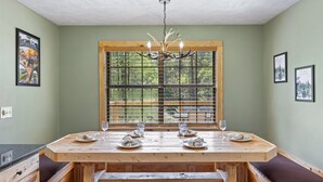 Comedor con una mesa de madera dispuesta para dos, una ventana con vistas a la vegetación y una araña.