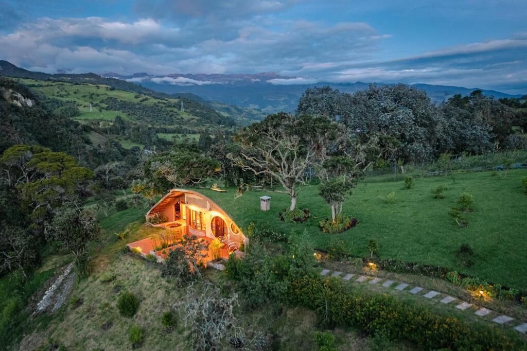 Una casa con techo curvo y ventanal circular, rodeada de un paisaje verde exuberante y montañas distantes.