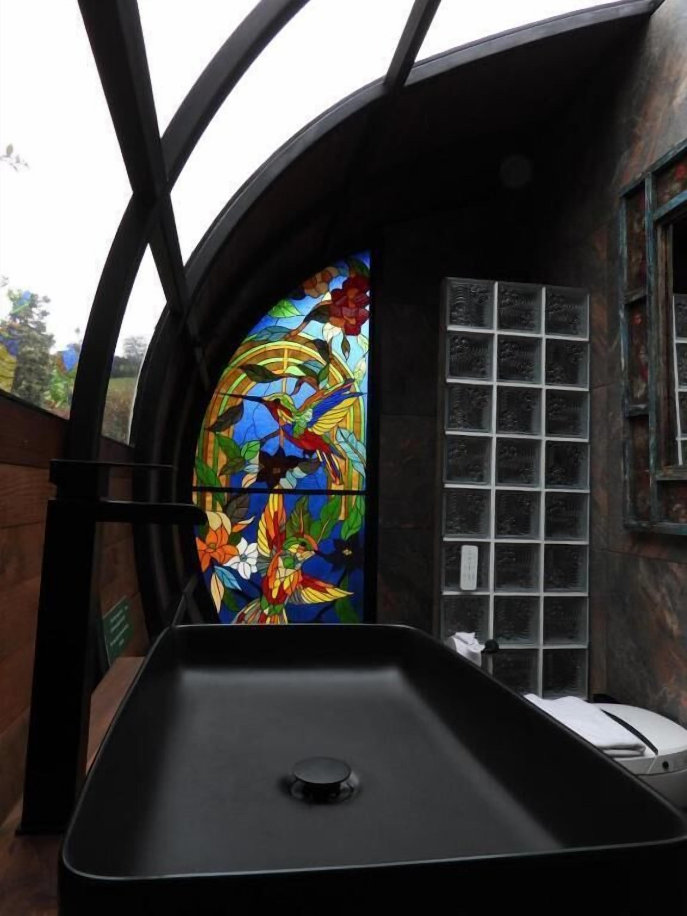 Een kamer met een groot rond glas-in-loodraam met vogels en bloemen, een zwarte wastafel en uitzicht op het buiten.