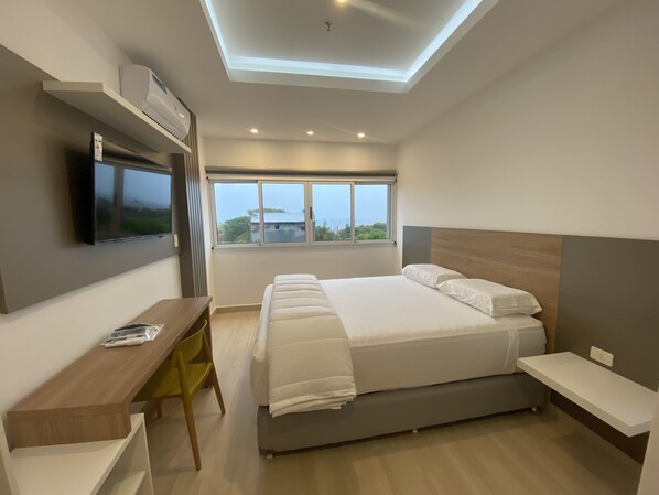 A modern bedroom with a large bed, a desk, a TV mounted on the wall, and a window with a view of greenery and buildings. - 25homeapart (Encarnacion)