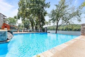 A swimming pool with a view of a lake and trees, surrounded by a building with a brick wall and lounge chairs.