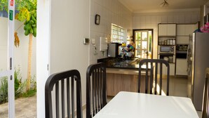 A modern kitchen with white cabinets, a dining area with a white table and black chairs, and a view of a tropical garden outside. - Hostel Santa Rita (Santa Rita De Cassia)