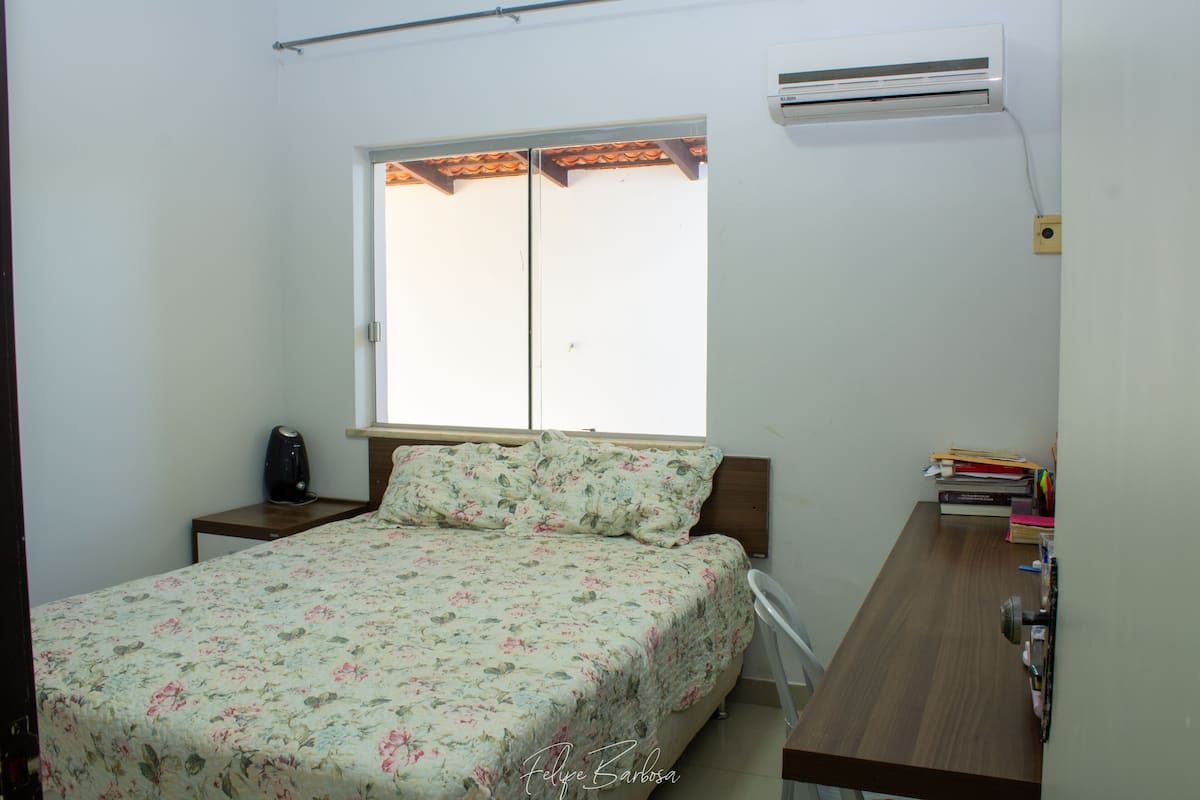 A bedroom with a bed, a nightstand, a desk, and an air conditioning unit.