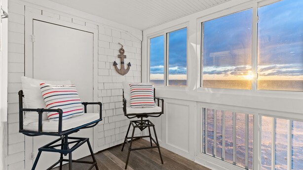 Ein Zimmer mit zwei schwarzen Metallstühlen mit weißen Kissen und gestreiften Kopfkissen, einer weißen Ziegelwand und einem großen Fenster mit Blick auf den Sonnenuntergang über dem Meer.