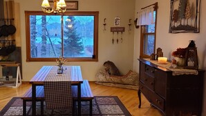 A cozy kitchen with a large window, wooden table, and various kitchen utensils hanging on the wall.