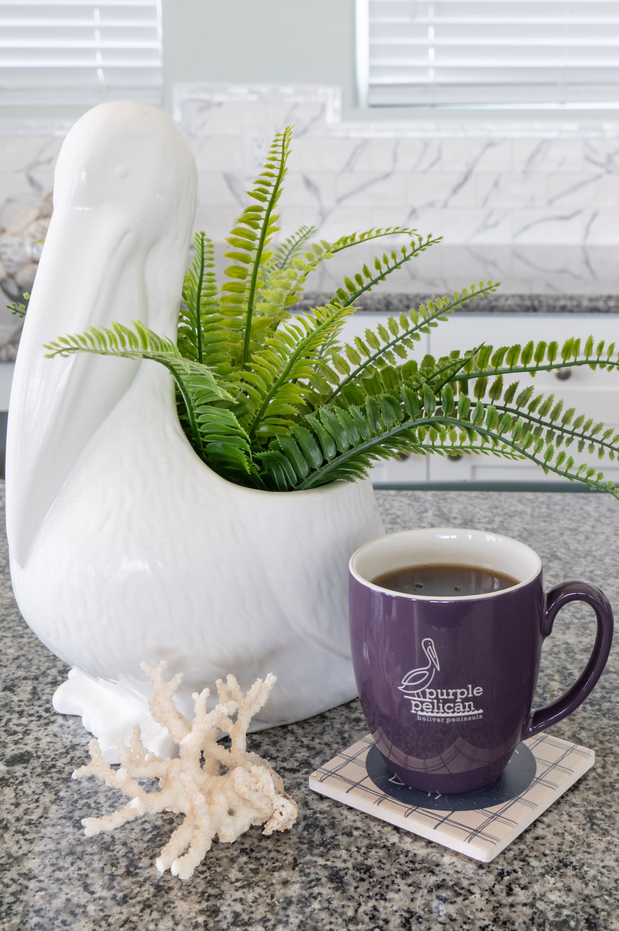 Un cisne de cerámica blanca sosteniendo un helecho, una taza morada con el logo de un pelícano y un adorno coral sobre una encimera de cocina.