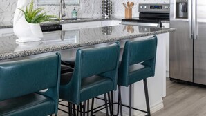 A modern kitchen with white cabinets, a granite countertop, a stainless steel refrigerator, and teal bar stools. - Beachfront • Perfect for Groups (Bolivar Peninsula)