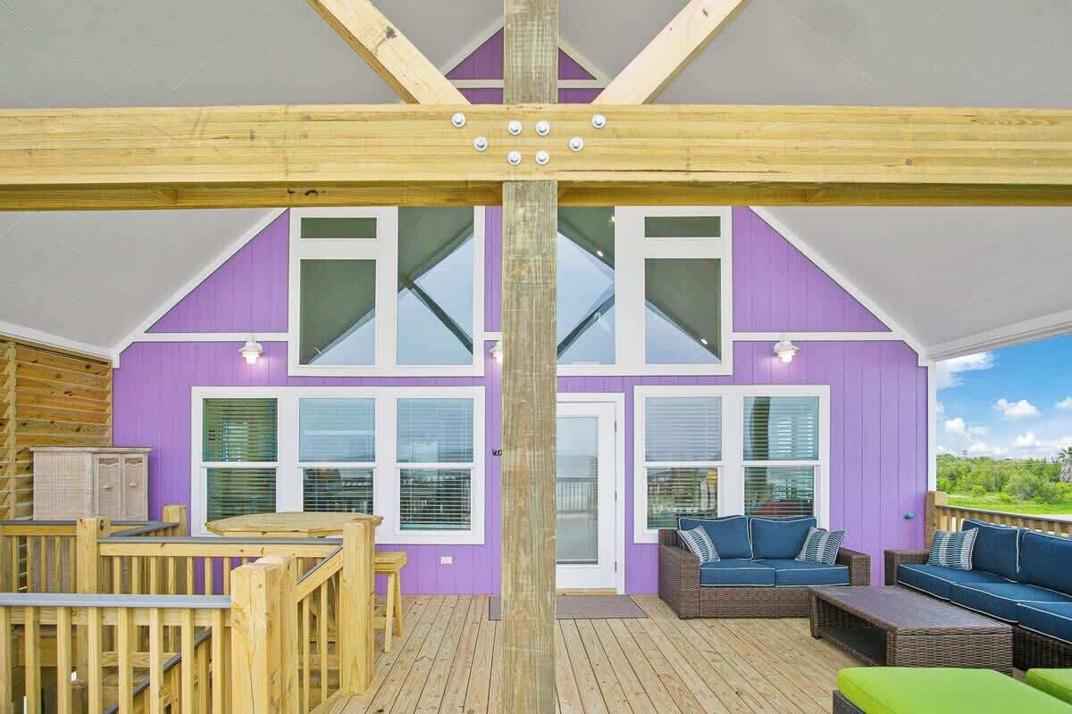 Une terrasse en bois avec une rambarde en bois, un mur extérieur de couleur violette et une grande fenêtre.