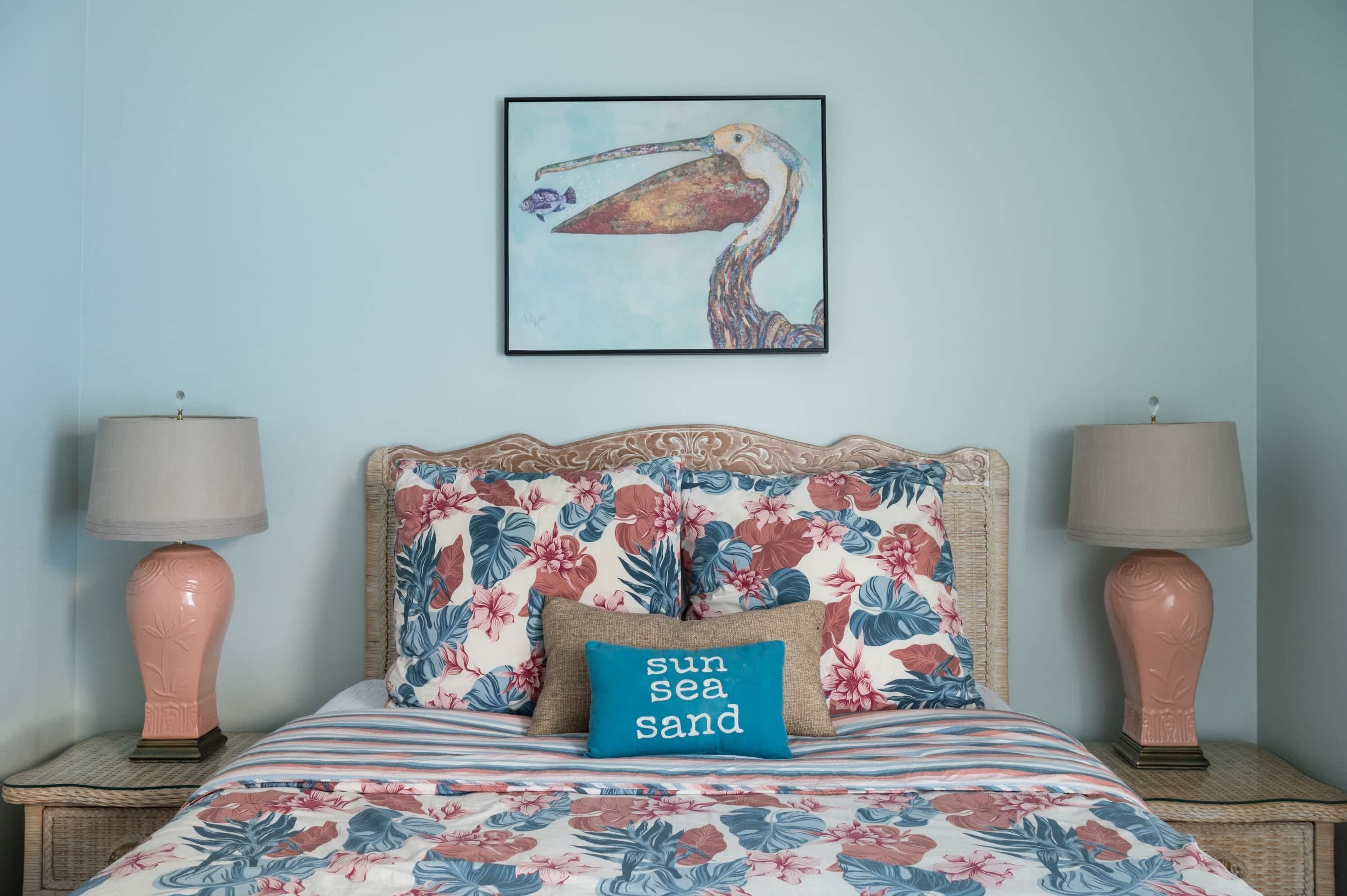 A bed with floral bedding, a pillow with "sun sea sand" text, and a painting of a pelican on the wall.