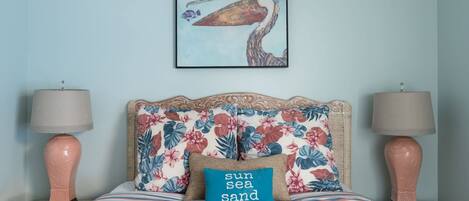 A bed with floral bedding, a pillow with "sun sea sand" text, and a painting of a pelican on the wall.