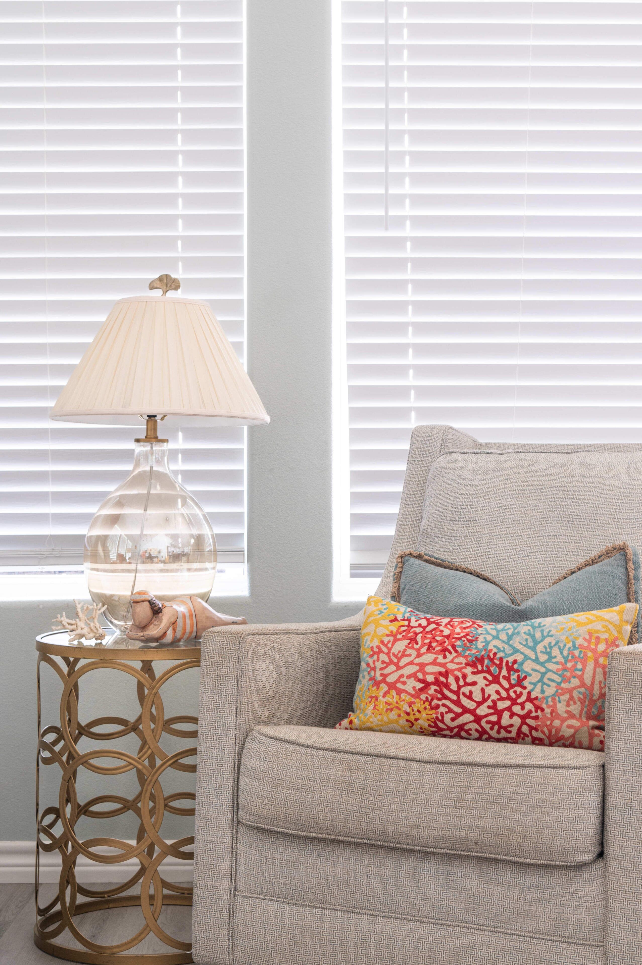 A room with a beige armchair, a colorful patterned pillow, a clear glass lamp, and a gold-rimmed side table.
