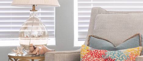 A room with a beige armchair, a colorful patterned pillow, a clear glass lamp, and a gold-rimmed side table.