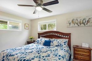Une chambre à coucher comprenant un lit, un ventilateur de plafond, deux fenêtres, une table de chevet, une commode et une décoration murale représentant des oiseaux.