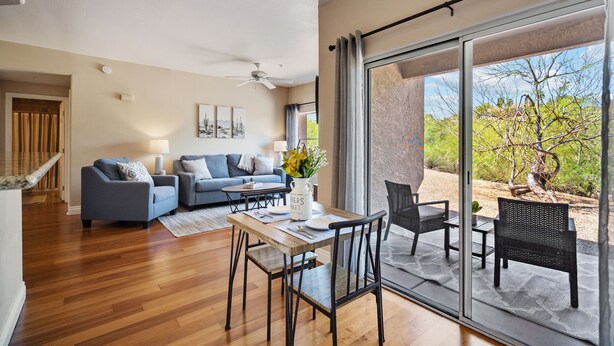 A modern living room with a dining area, a ceiling fan, and a large sliding glass door leading to an outdoor patio with a view of trees and a desert landscape. - Enjoy Paradise in this Quiet, Resort-Style 2 BR Condo (Fountain Hills)