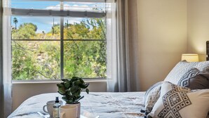 A bedroom with a large window, a bed with patterned pillows, a bedside table with a plant and a bottle, and a lamp on the nightstand. - Enjoy Paradise in this Quiet, Resort-Style 2 BR Condo (Fountain Hills)