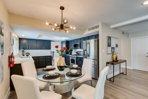 Una cocina moderna con gabinetes oscuros, electrodomésticos de acero inoxidable y una mesa de comedor de vidrio con sillas blancas.