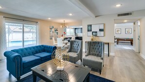 A spacious living room with a blue sofa, grey armchairs, and a coffee table with a decorative object. There is a dining area in the background with a pool table.