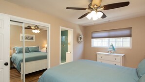 Un dormitorio con una cama, ventilador de techo, ventana con persianas, mesita de noche y un espejo.