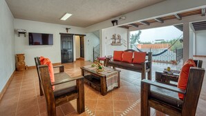 A spacious living area with a wooden coffee table, two sofas with orange cushions, a flat-screen TV, and a balcony with a view of the city.