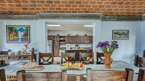 A dining area with a wooden table, chairs, and a vase of flowers. There is a kitchen in the background with wooden cabinets and a refrigerator.