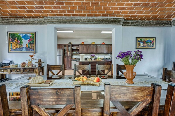 Un comedor con una mesa de madera, sillas y un jarrón de flores. Se observa una cocina al fondo con armarios de madera y un refrigerador.