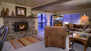 A cozy living room with a stone fireplace, a leather armchair, a wooden coffee table with a lamp, and a view of the outdoors through a sliding glass door. - Evergreen Condo | Walk to Lifts | Shared Hot Tub | Very Convinient (Mt. Crested Butte)