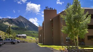 A parking lot with several vehicles, a building with a flat roof and multiple chimneys, and a mountain in the background. - Evergreen Condo | Walk to Lifts | Shared Hot Tub | Very Convinient (Mt. Crested Butte)
