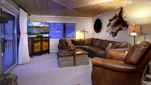 A living room with a brown leather sofa, a television, a mounted animal hide, and a stone fireplace. - Evergreen Condo | Walk to Lifts | Shared Hot Tub | Very Convinient (Mt. Crested Butte)