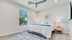 Un dormitorio con cama, mesitas de noche, ventilador de techo, ventana con vista a árboles y televisor montado en la pared.