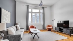 Uma sala de estar moderna com um sofá cinzento, uma mesa pequena, duas poltronas, uma televisão e uma janela grande com cortinas.