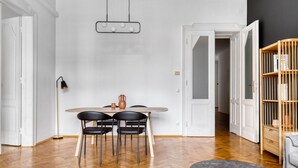 Uma sala de estar moderna com piso de madeira em padrão herringbone, uma mesa de jantar redonda com duas cadeiras pretas, uma luminária pendente e uma estante com livros.