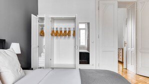 Quarto com cama, guarda-roupa cheio de roupas e vista para outro cômodo.
