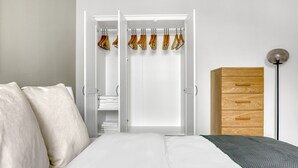 A bedroom with a bed, a wardrobe with hanging clothes, and a wooden dresser.