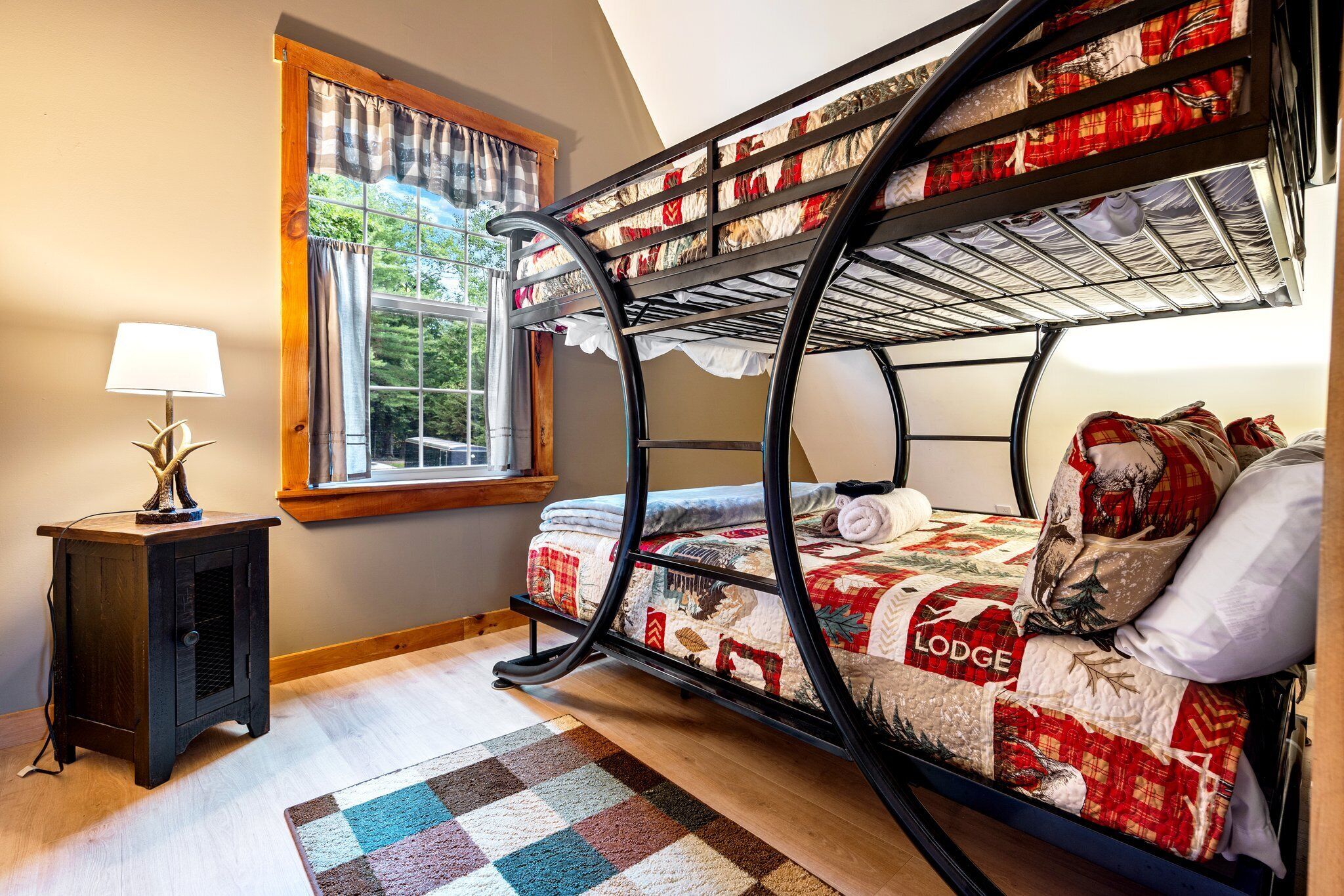 Een stapelbed met een gewatteerde spreidekbed, een klein houten nachtkastje met een lamp, en een raam met gordijnen.