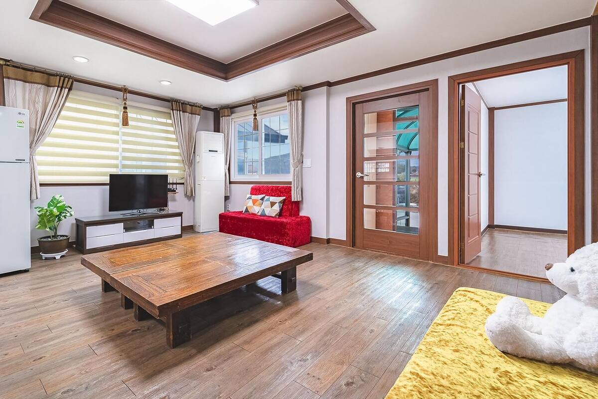 A modern living room with a wooden table, a red sofa, a TV, and a large window with blinds.