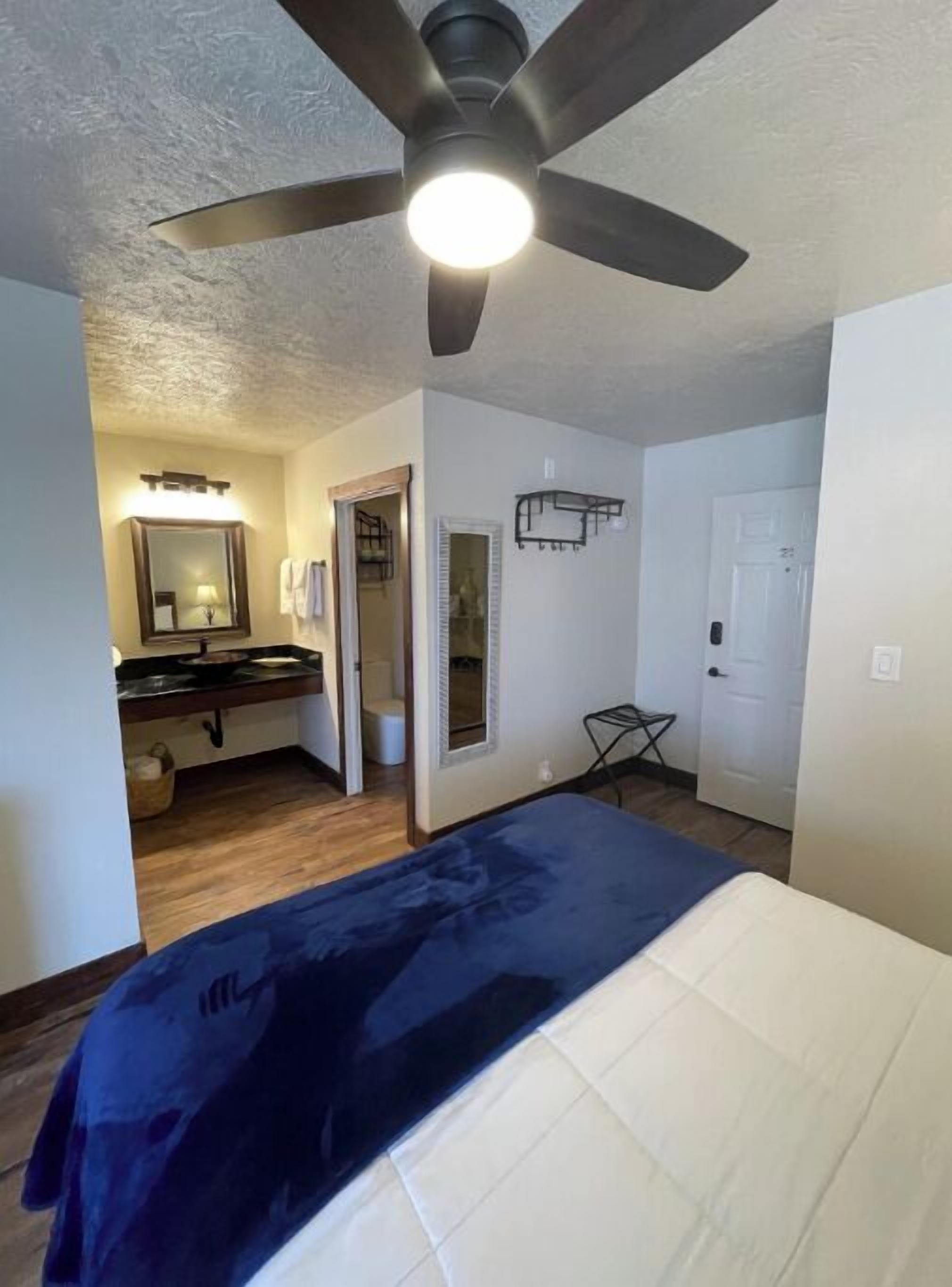 Une chambre à coucher avec un lit, un ventilateur de plafond et une salle de bain visible à travers une porte ouverte.