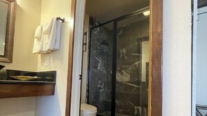 Un baño con piso de madera, inodoro blanco, cabina de ducha con vidrio y un lavamanos con espejo.