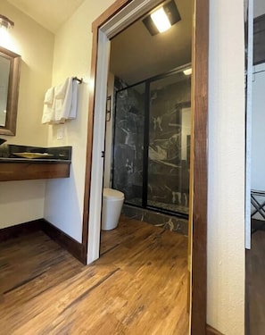 Une salle de bain avec un plancher en bois, des toilettes blanches, une douche en verre et un lavabo avec un miroir.