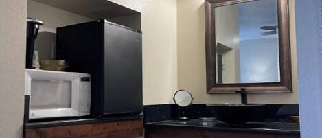 A bathroom with a white sink, a mirror, a microwave, and a dark wooden vanity.