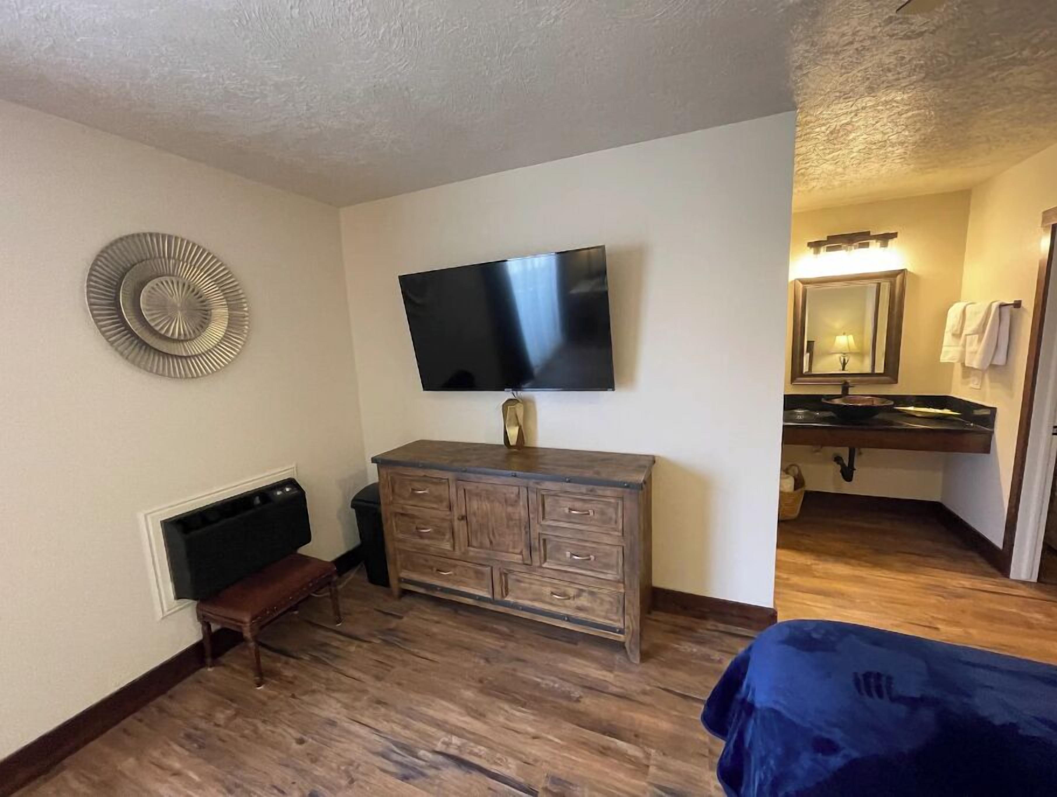 A room with a wooden dresser, a flat-screen TV mounted on the wall, a bench, and a bathroom with a sink and mirror.