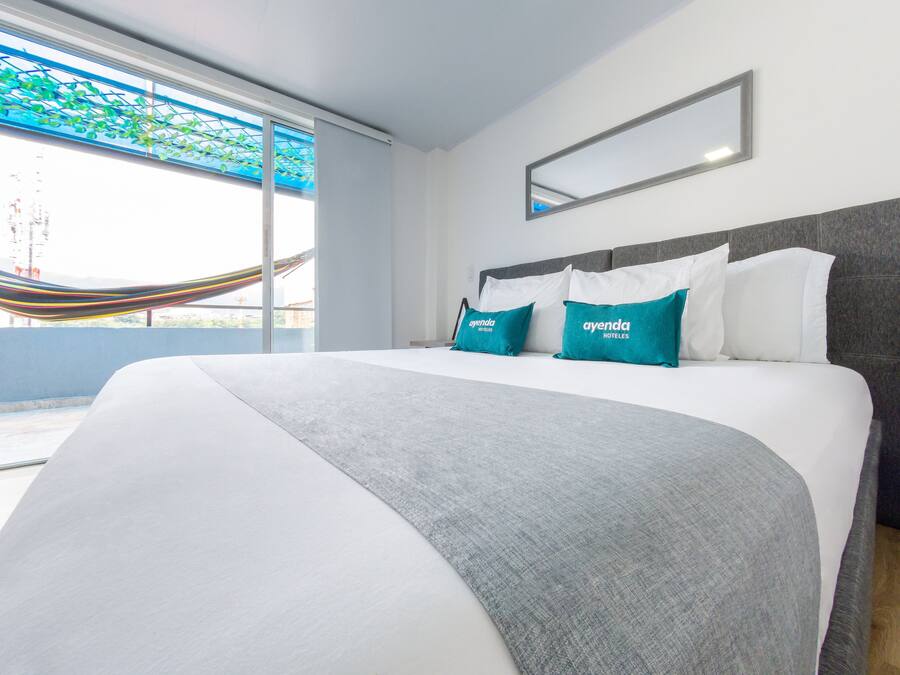 A modern hotel room with a large bed, a view of the city through a large window, and a hammock outside.