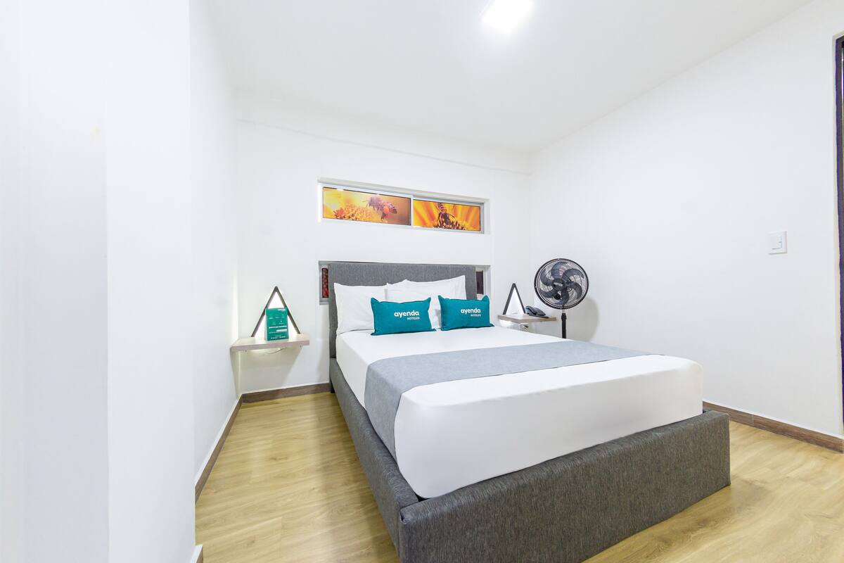 A hotel room with a bed, a nightstand, a fan, and a window with artwork.