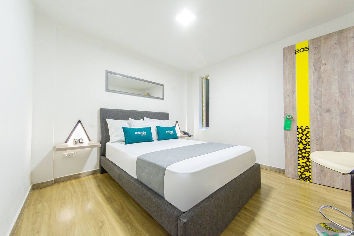 A modern hotel room with a large bed, a bedside table, a mirror, and a wooden door with a yellow stripe.