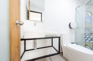 A bathroom with a wooden door, a white sink with a glass shelf, a towel, a mirror, a shower with patterned tiles, and a toilet paper dispenser.